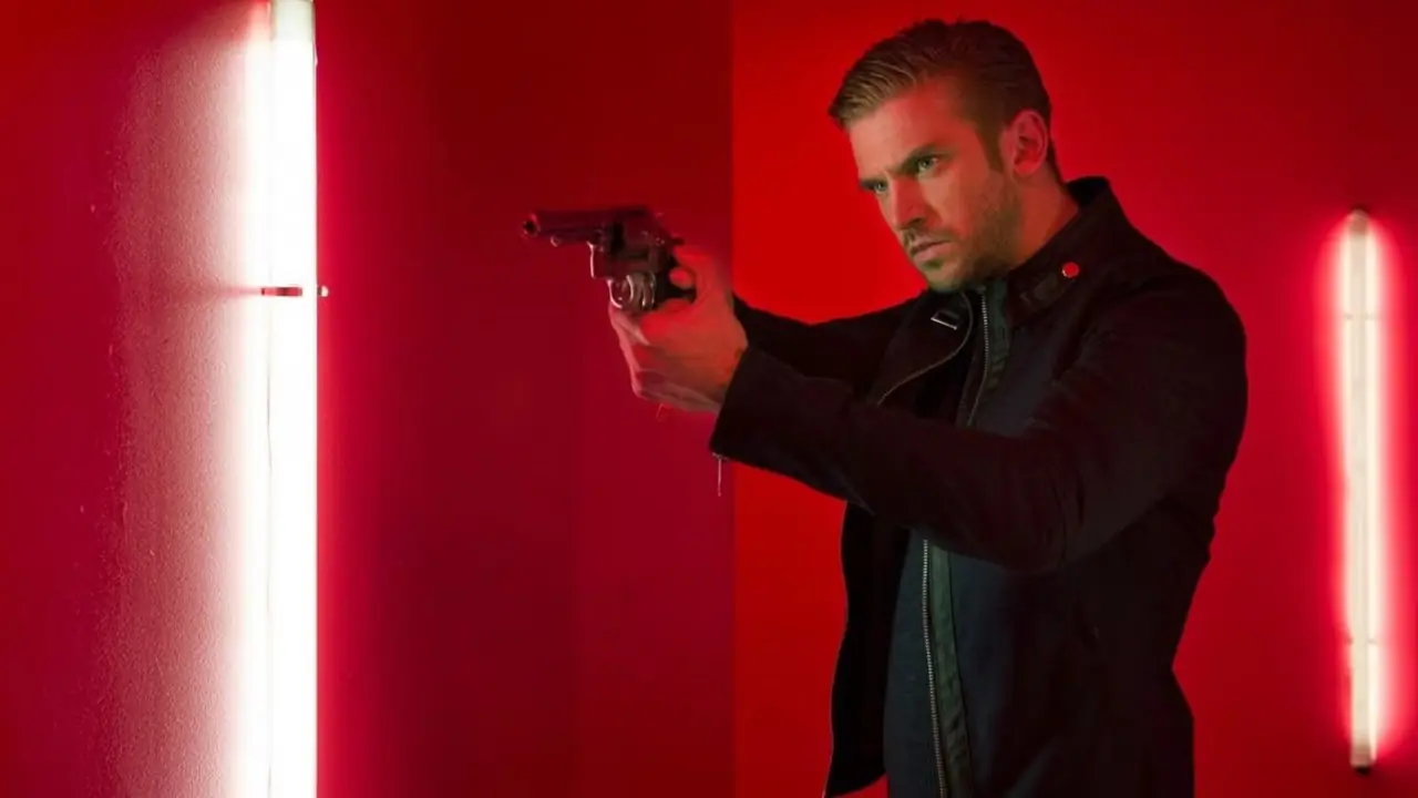 Dan Stevens apuntando con un revólver sobre un fondo rojo en The Guest Dan Stevens apuntando con un revólver sobre un fondo rojo en The Guest