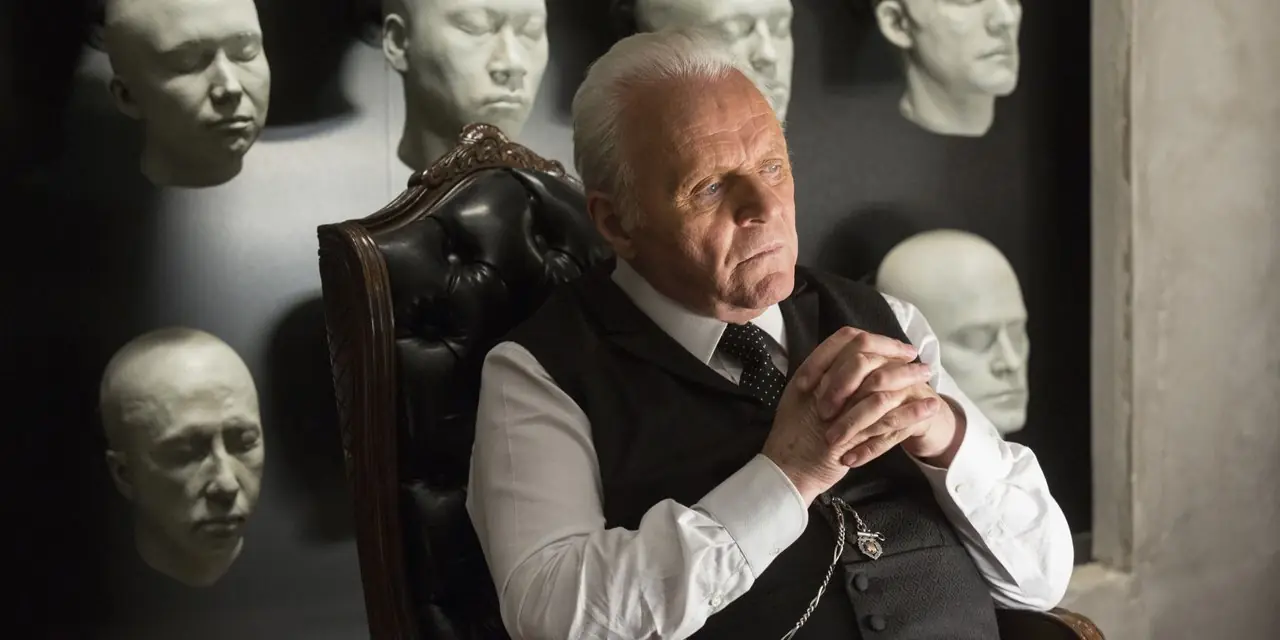 Anthony Hopkins como el Dr. Robert Ford en Westworld, sumido en sus pensamientos, sentado en una silla, con moldes de cabezas de los anfitriones del parque colgados en la pared detrás de él.