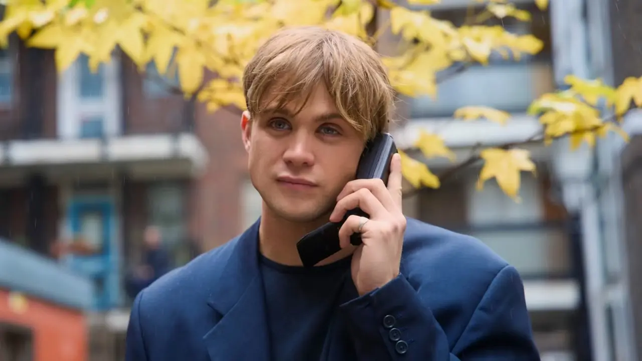 Dex uśmiecha się, rozmawiając przez telefon i stojąc przed drzewem o żółtych liściach w „Jeden dzień”. Dex uśmiecha się, rozmawiając przez telefon i stojąc przed drzewem o żółtych liściach w „Jeden dzień”.