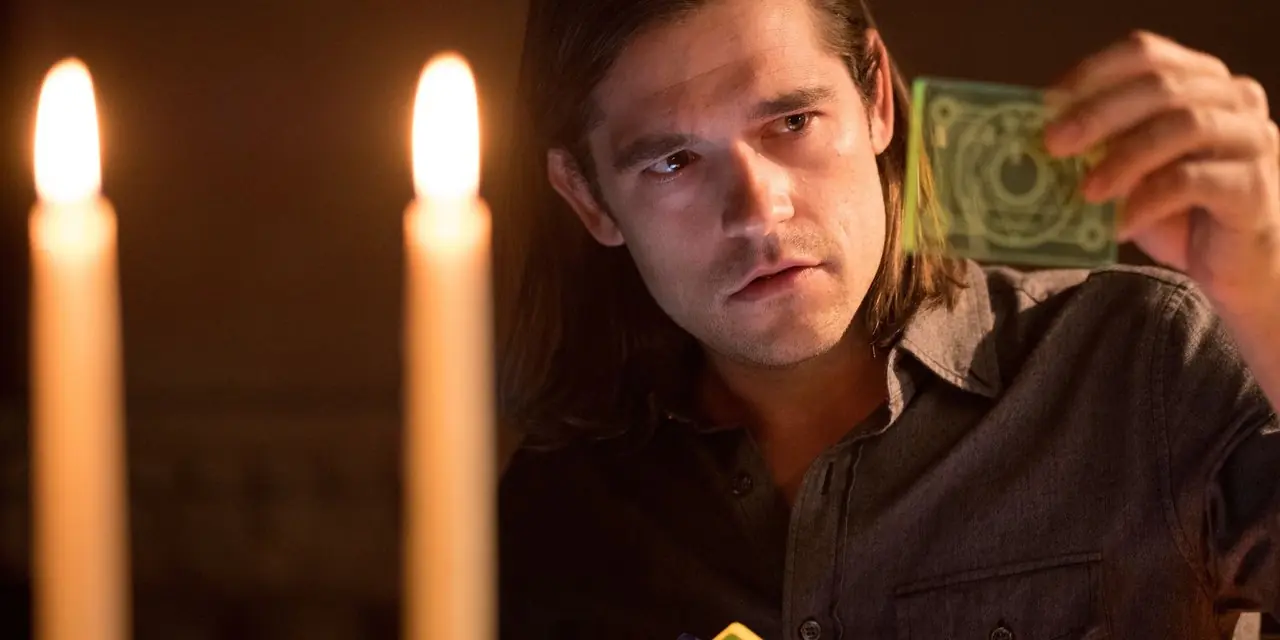 Jason Ralph como Quentin Coldwater sosteniendo una carta mágica con dos velas frente a él en Los Magos