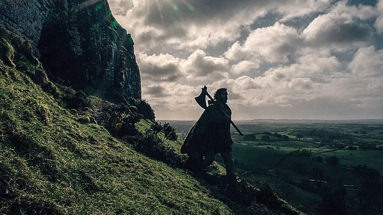 Gawain (Dev Patel) caminando cuesta abajo, llevando un hacha en The Green Knight.