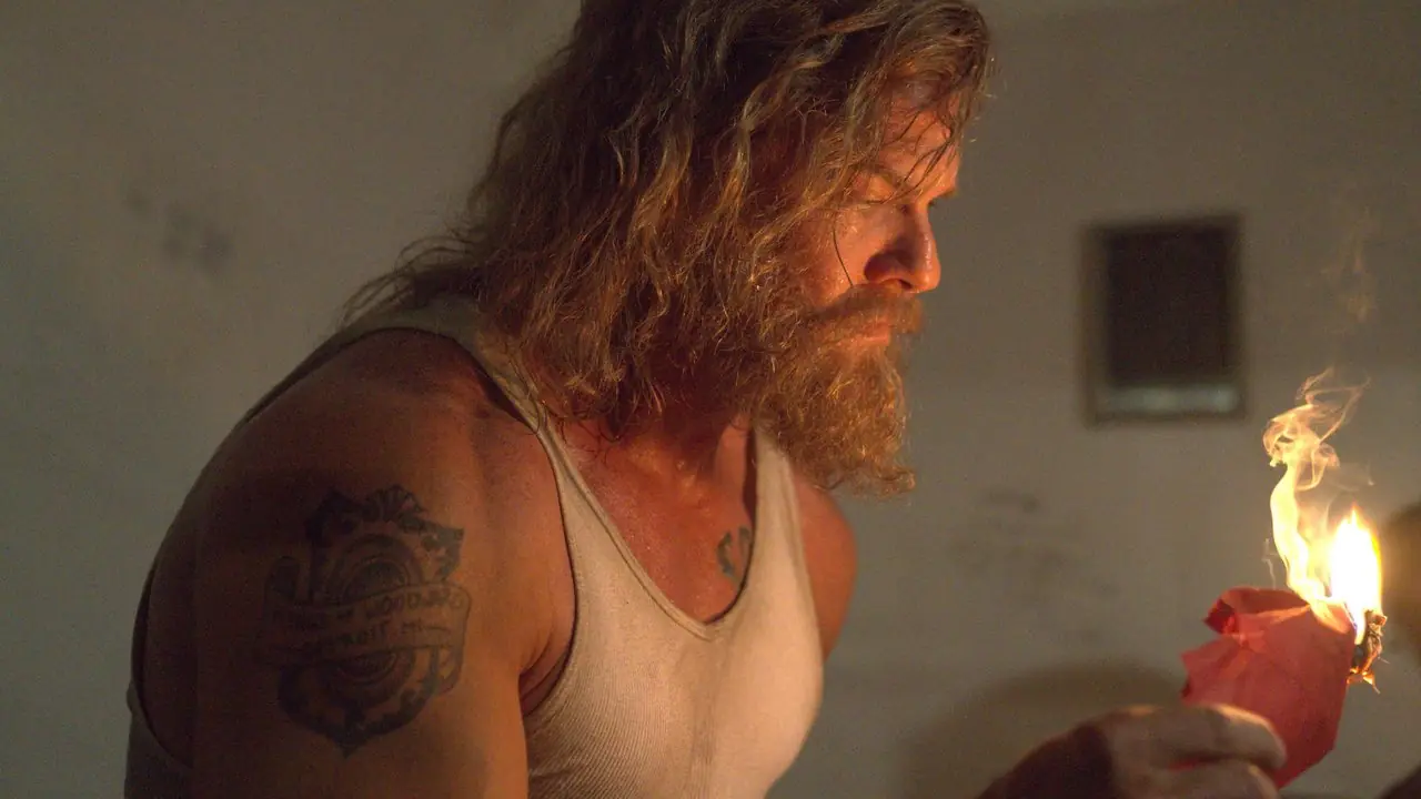 Alan Ritchson con i capelli lunghi e la barba che brucia un giornale a Motor City