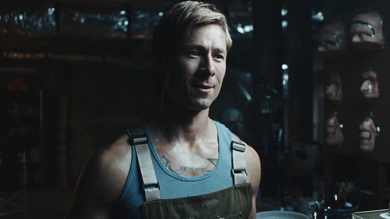 चाड पॉवर्स सीज़न 2 में ग्लेन पॉवेल एक तहखाने में दीवार पर अपने चारों ओर मुखौटे के साथ